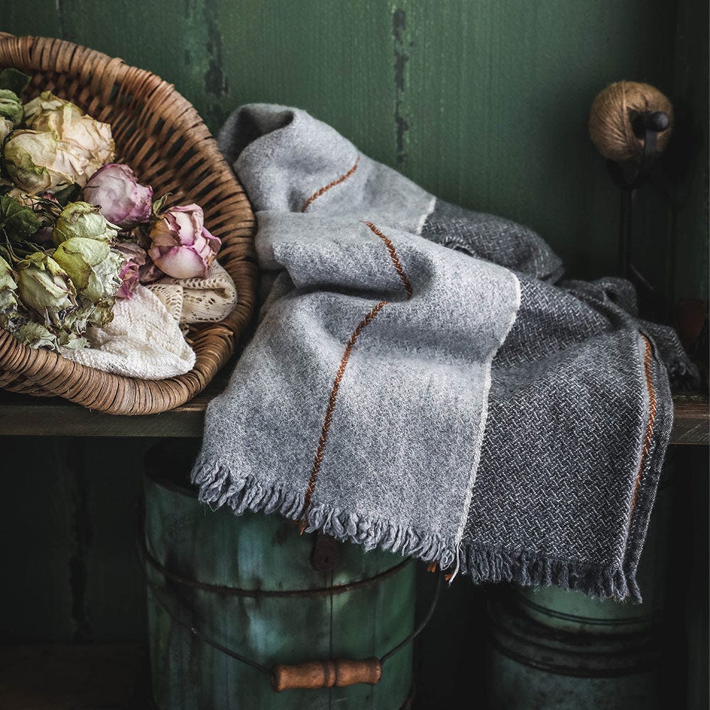A gray striped throw draped on a wood bench sits next to a wooden basket filled with dry florals.
