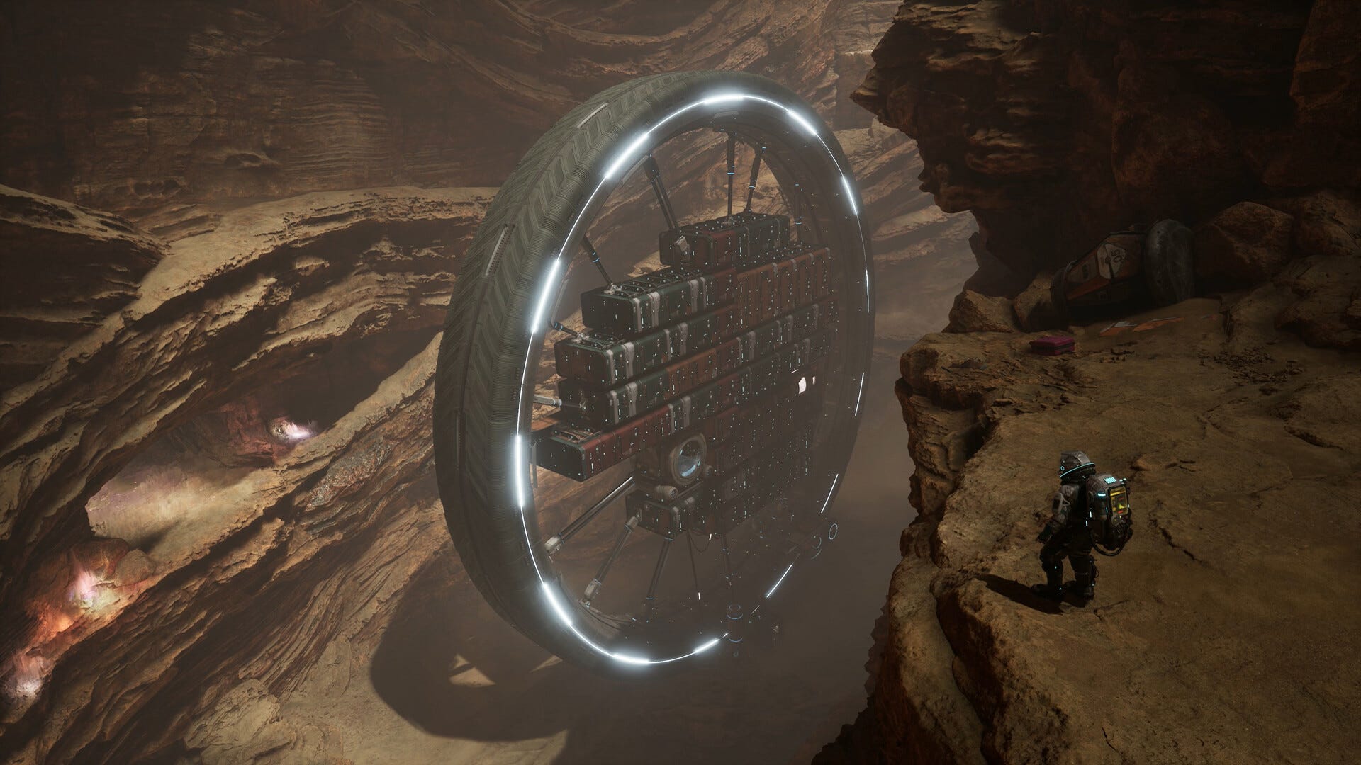 Video game screenshot of a man in a spacesuit, standing on a rocky cliff. In the valley in front of him is a massive wheel-shaped based.