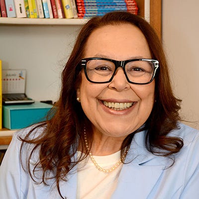 Mulher branca, cabelos e olhos castanhos. Usa óculos de armação escura, blazer azul caro e blusa branca. Fundo, estante de livros.