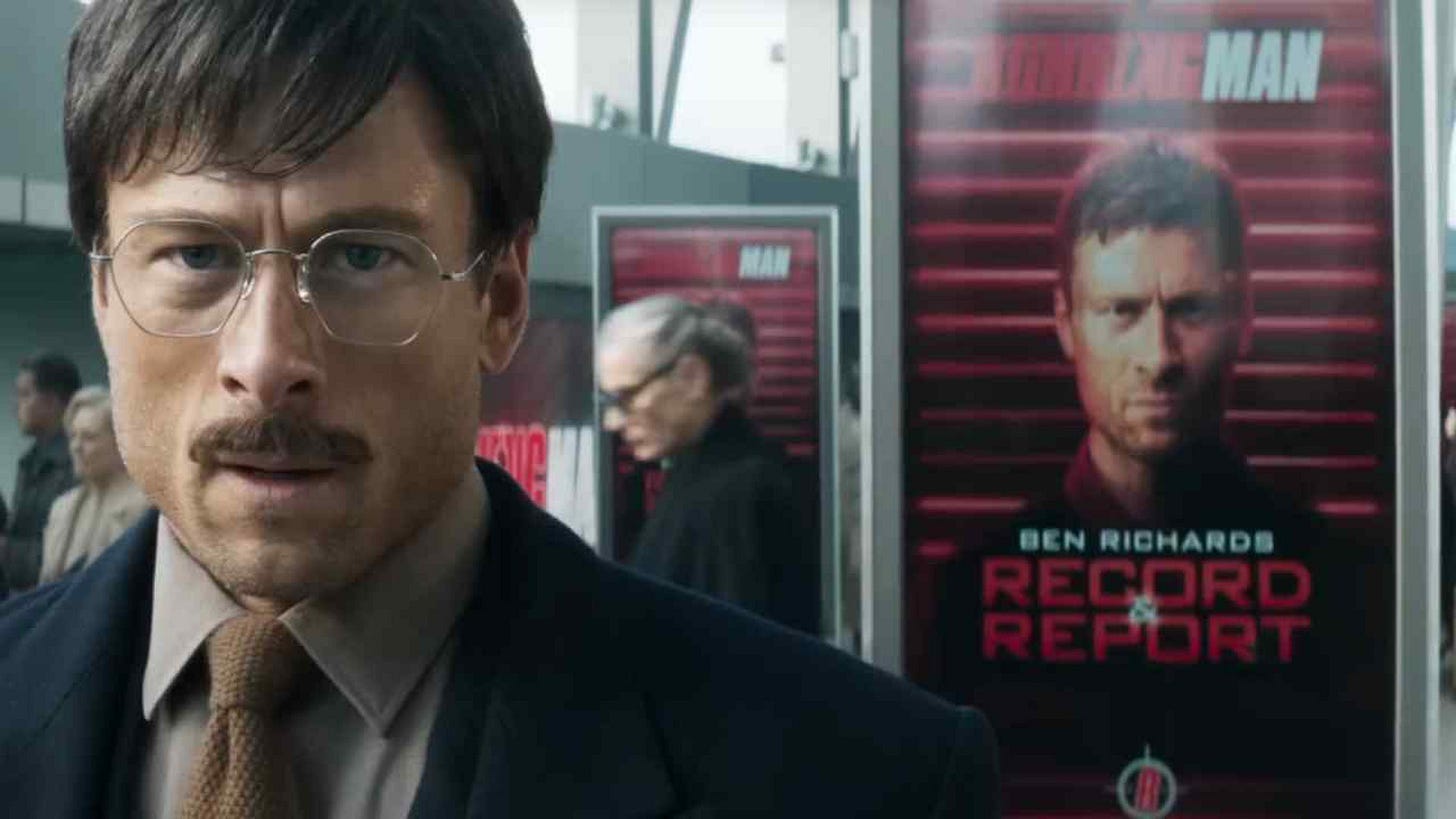 still from Running Man; Powell with glasses and glasses stands in front of a screen showing him without those and advertising the Running Man show within a show