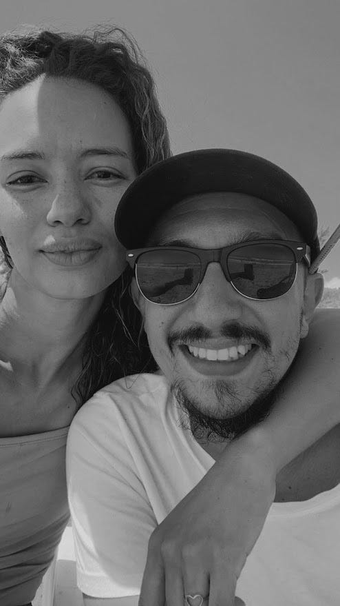 mulher linda, cachos longos ao vento, sorriso com os lábios grossos fechados. homem branco de óculos escuros, boné preto, bigode e cavanhaque sorrindo para a foto. foto em preto e branco.