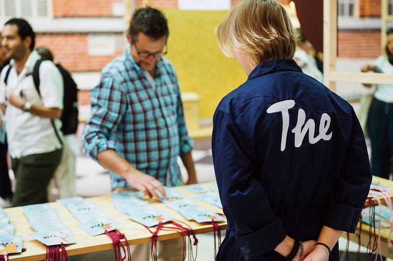 A volunteer at the Swedish stands with their hands clasped behind their back at the ideas conference called The Conference in their trademark navy blue overalls with a stylised white "The" logo on its back. The volunteer is at the delegate check-in table and is assisting a man find his name badge.