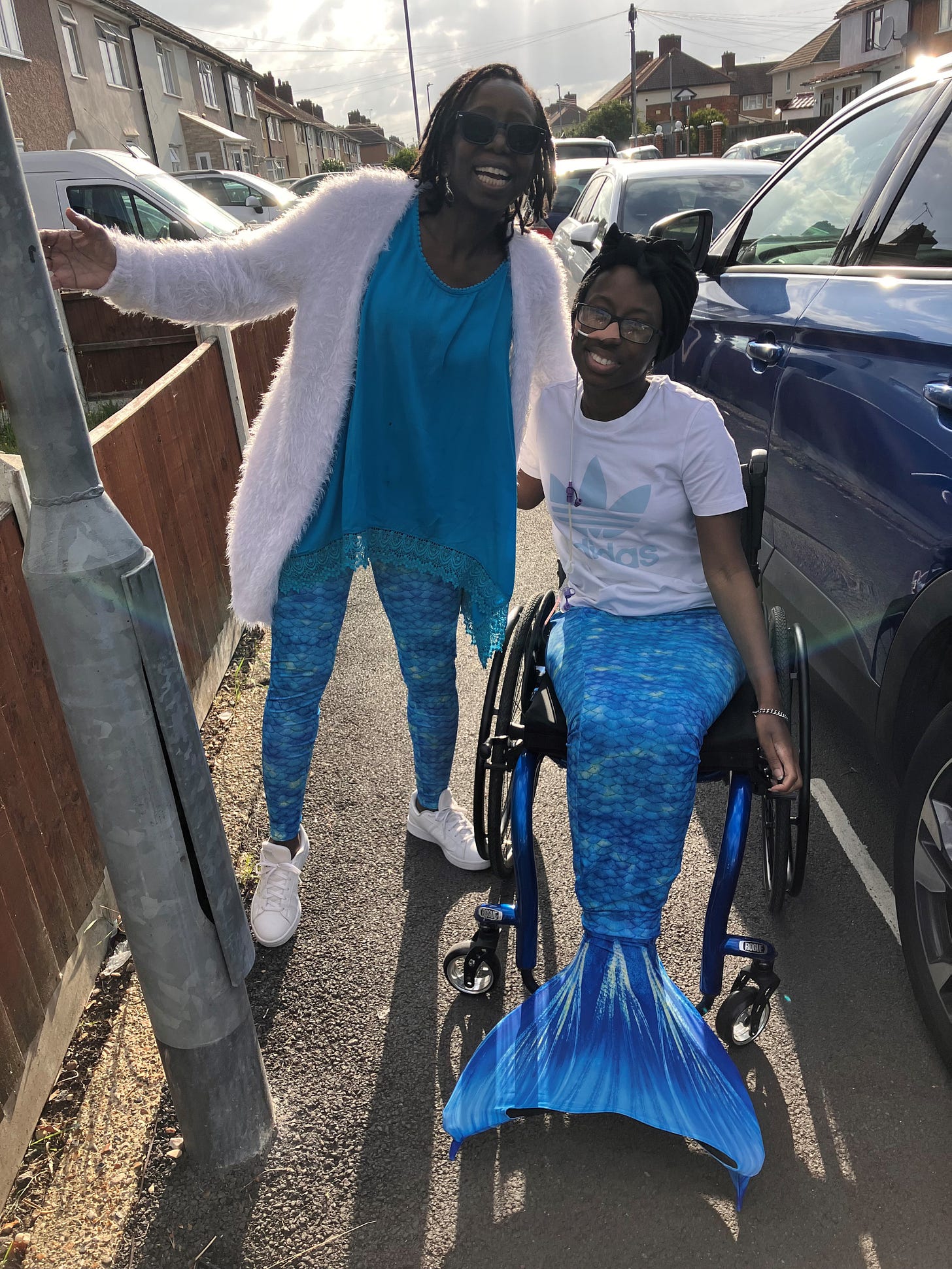 Cyrene is with her mum — a Black woman wearing leggings with the same mermaid print as Cyrene’s mermaid tail, with a bright blue top, a white cardigan, and white trainers. She is also wearing sunglasses. She is next to Cyrene with one arm around her and the other arm up to the side of her in an enthusiastic stance. Cyrene has one hand leaning on her wheelchair seat/brake handle, and one hand around her mum. Both of them have big smiles on their faces. 