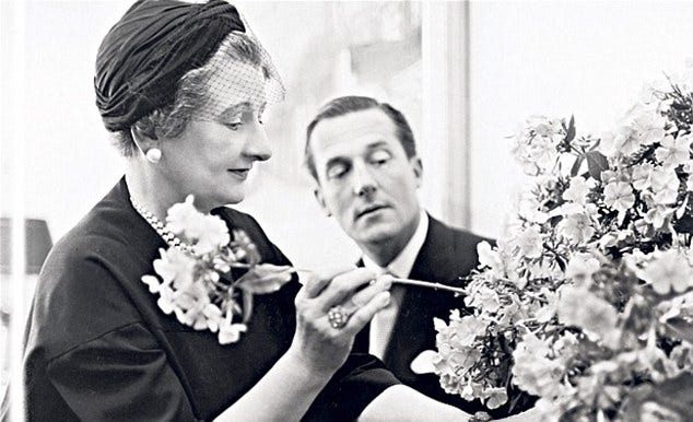 1) Constance is a white woman in a dark outfit and hat with lace coming off the back holding a sculpted vase shaped like a face with roses sticking out of the top, her expression curious and intrigued, against a plain backdrop. The vase sits on a ridged pillar, and she is examining the flower arrangement to see if she's happy with how it looks. She has on a small hoop earring, and is wearing rings on her left ring finger. 2) A black-and-white photo of a white woman in a head scarf arranging flowers, with a white man observing her work. The setting feels elegant and focused. Constance is arranging a floral bouquet, and she is holding a flower that she is about to place. She has on a pearl necklace, and earring, and she has rings on her right ring finger. She is wearing dark clothing, and the man watching behind her is wearing a suit.