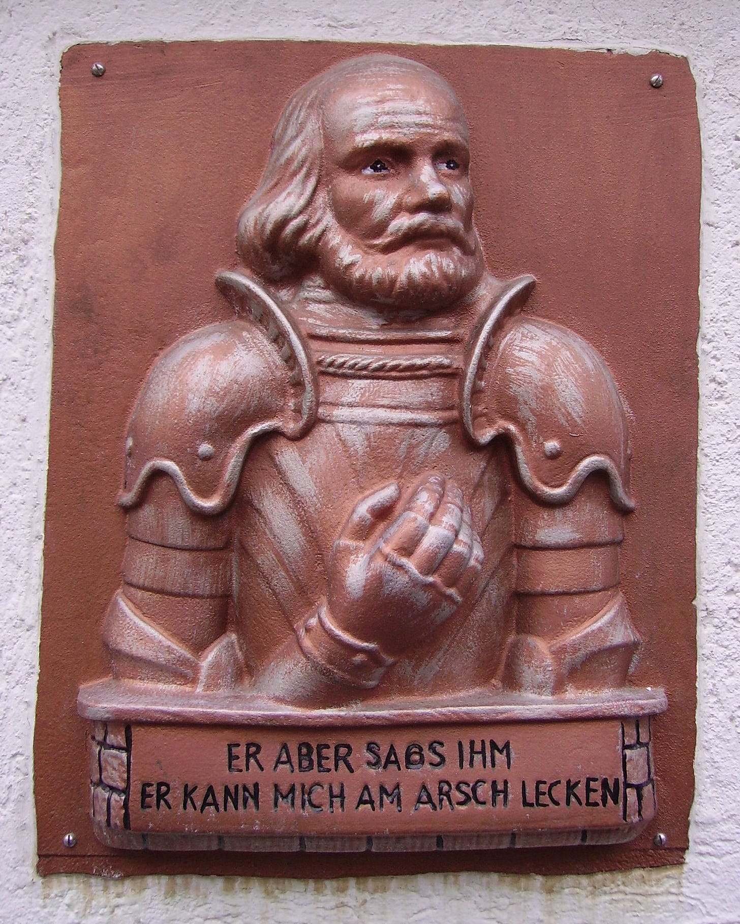 A brass effigy of Götz wearing his armor. He has a receding hairline with long blond locks and a full beard. He holds his iron hand over his chest so the mailed fist is visible. The accompanying text reads “ER ABER SABS IHM ER KANN MICH AM ARSCH LECKEN.” TRANSLATION: And as for him, he can lick my ass. A brass effigy of Götz wearing his armor. He has a receding hairline with long blond locks and a full beard. He holds his iron hand over his chest so the mailed fist is visible. The accompanying text reads “ER ABER SABS IHM ER KANN MICH AM ARSCH LECKEN.” TRANSLATION: And as for him, he can lick my ass.
