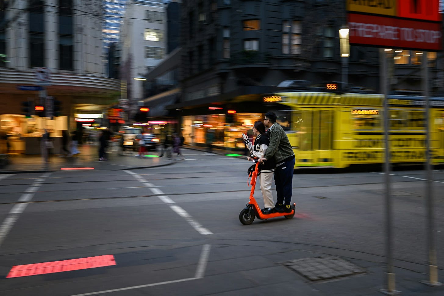 Two people ride together on an orange electric scooter through a busy intersection in Melbourne’s CBD at dusk, with a yellow tram and blurred shopfronts in the background.