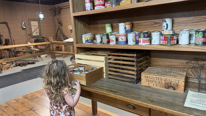 A curly-haired girl picking out cans of vegetables from an interactive mercantile. | Close up of black, lace-up ladies boots. | Low angle from behind the sales counter showing off several cupboards and an old cash register. | Same girl checking mail in the postal cubbies.