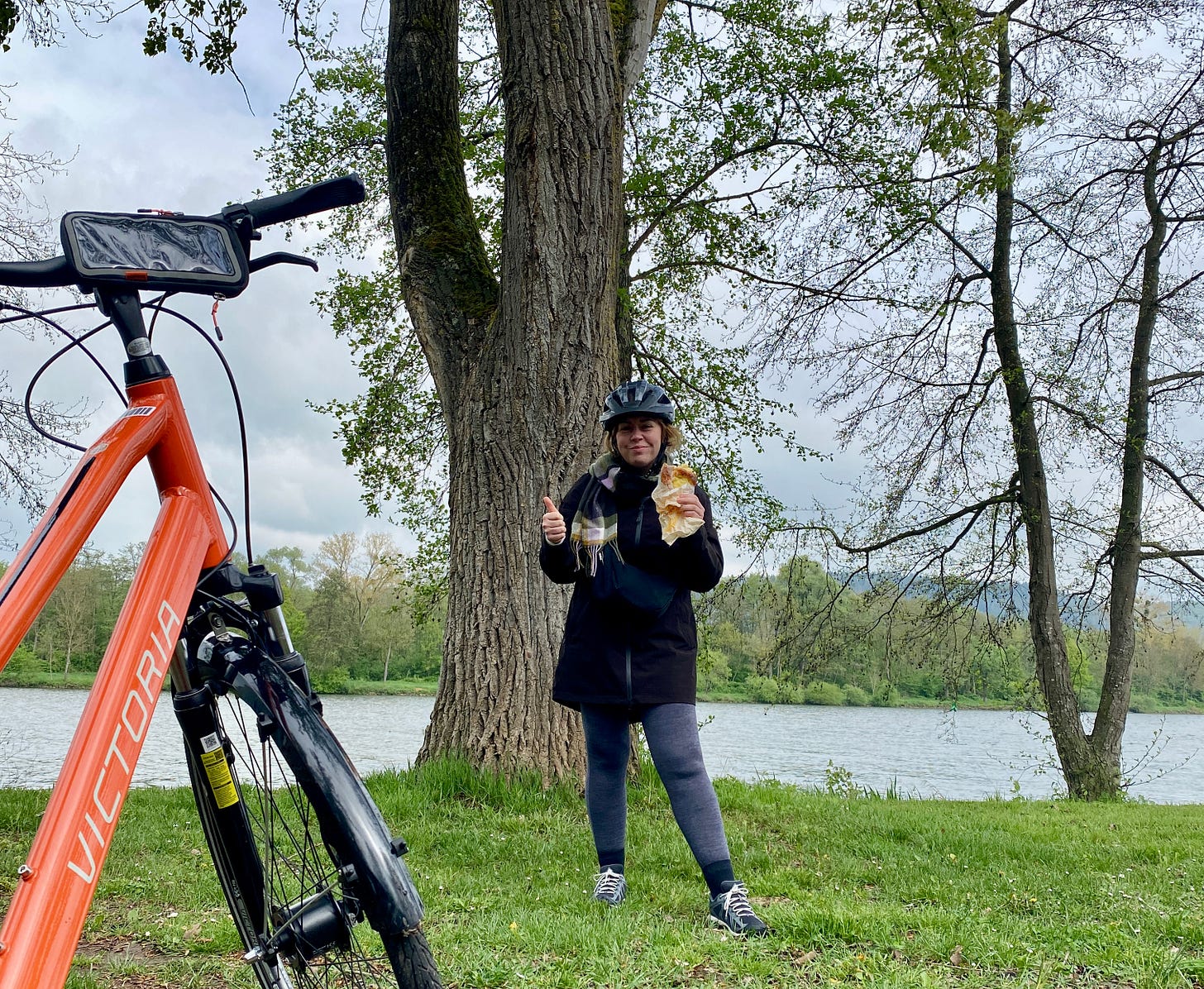 A woman holding a pastry and her thumb up as she smiles wearing a helmet and her bike in the foreground