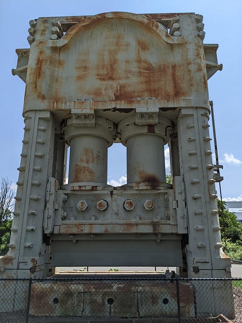 The remains of a massive 12,000 ton press now a rusting frame standing outdoors behind a chain-link fence on a bright blue day.