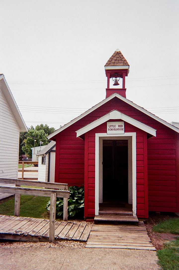 The exterior of a red schoolhouse. | The interior with a stovepipe furnace in the center of two rows of wooden desks.