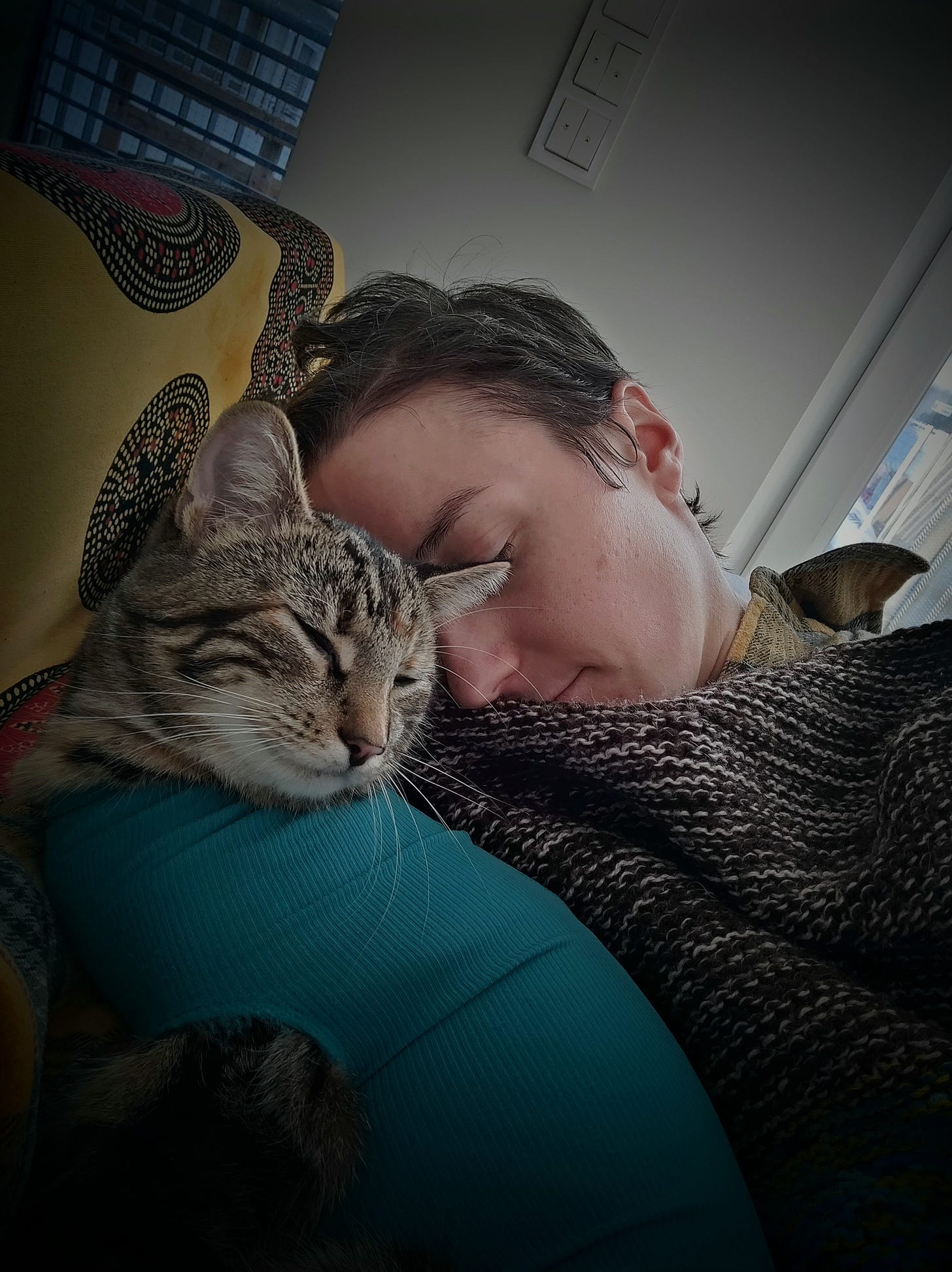 A white woman with short brown hair is sleeping on a couch with a cat in a turquoise onesie. A white woman with short brown hair is sleeping on a couch with a cat in a turquoise onesie.