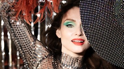 Sophie Ellis-Bextor wears a sequined outfit with dark burnt orange feathers, Only half her face is visible behind the brim of a black bejewelled hat she holds. She wears bright blue/green eyeshadow and bright red lipstick. Sophie Ellis-Bextor wears a sequined outfit with dark burnt orange feathers, Only half her face is visible behind the brim of a black bejewelled hat she holds. She wears bright blue/green eyeshadow and bright red lipstick.