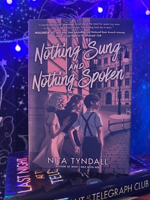 A book titled Nothing Sung and Nothing Spoken by Nita Tyndall stands upright against my glowing background with purple lights and constellation tapestries. The cover illustration shows three teenagers walking down a European-style street at dusk, with buildings and streetlights behind them. Text at the top reads: “A heartbreaking and bittersweet novel about the need for queer joy even in the midst of the horrors of war. The ending had me in tears.” —Malinda Lo, New York Times bestselling and National Book Award–winning author of Last Night at the Telegraph Club. Additional text reads: “NITA TYNDALL, author of Who I Was With Her.”