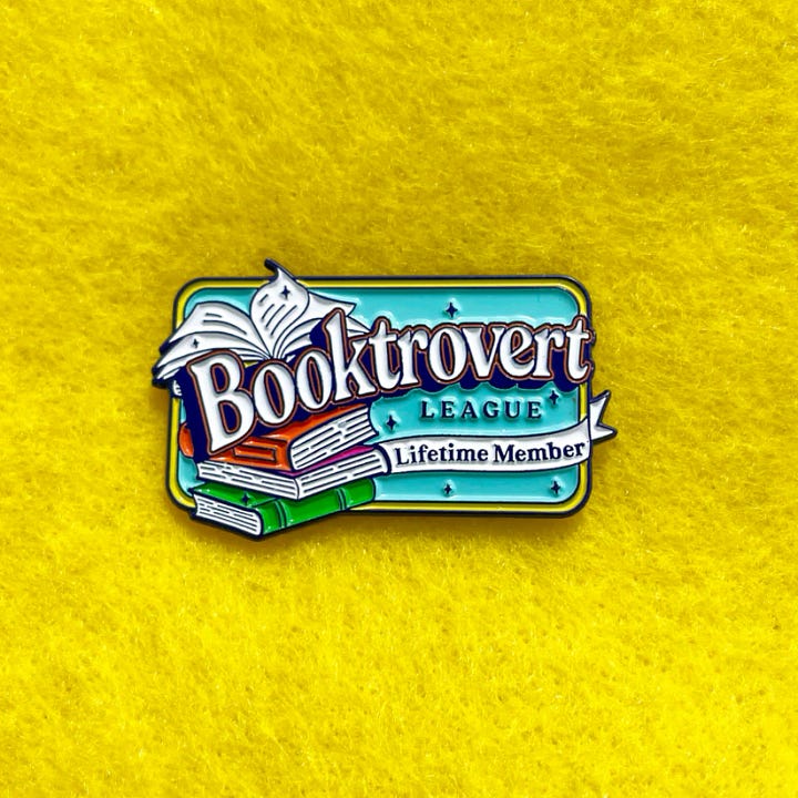 First photo: an enamel pin that says, "Booktrovert League: Lifetime Member."  Second Photo: a group of cat enamel pins in various designs that look like baked goods.