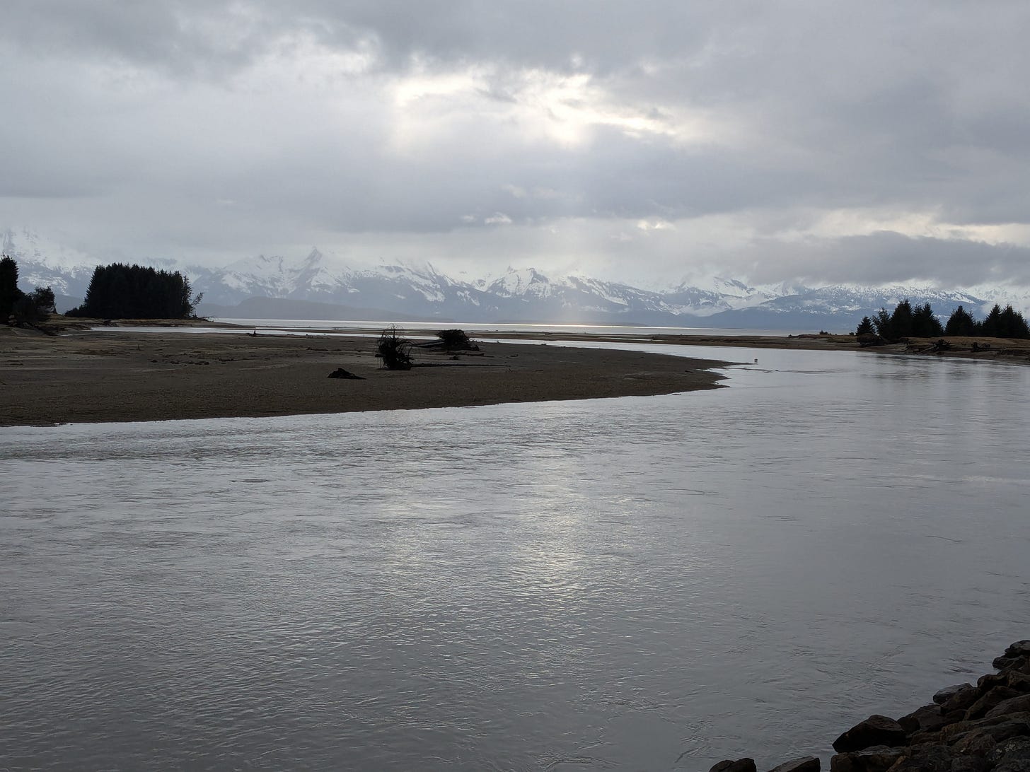 An overcast day near Eagle Beach in Alaska, with the river in the foreground. Two sunbeams are pouring out of the clouds in front of the mountain range in the distance.