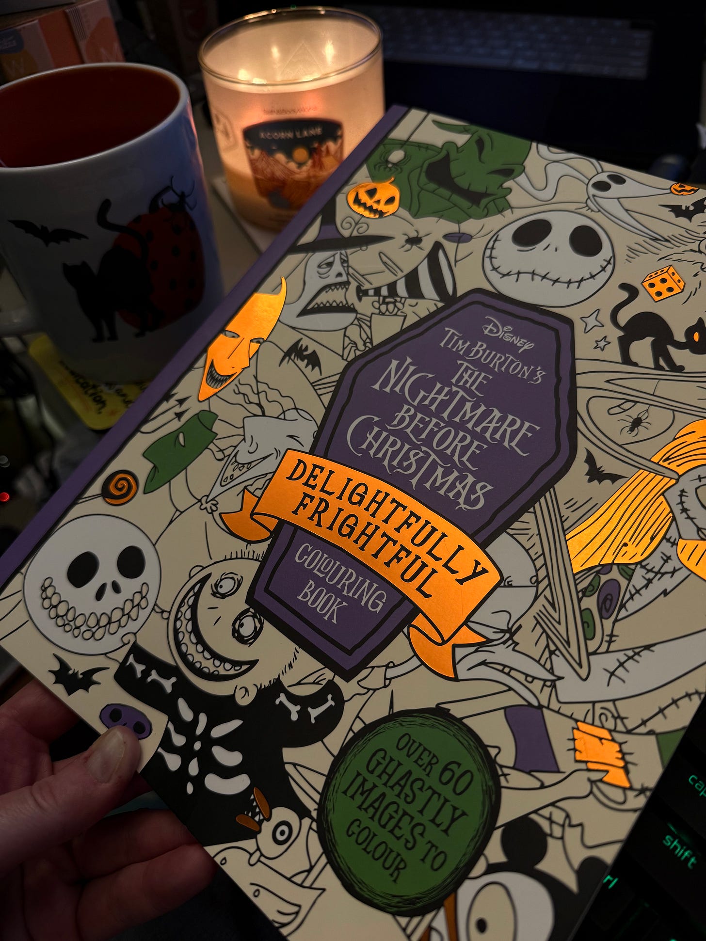 A hand holds up "The Nightmare Before Christmas: Delightfully Frightful" colouring book, with a lit candle in the background, symbolizing cozy, autumnal creative mischief.