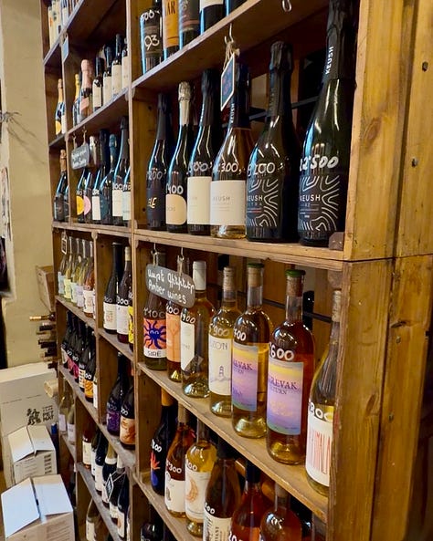 Photos inside In Vino wine bar in Yerevan, including a hand holding a bottle of Trinity Canyon Areni Noir, staff pouring wine behind shelves stocked with Armenian and international bottles, friends toasting with glasses of white wine, a long tasting table set with glassware beneath a sign reading 'The Renaissance of Armenian Wine Making,' and a wall of photos honoring winemakers