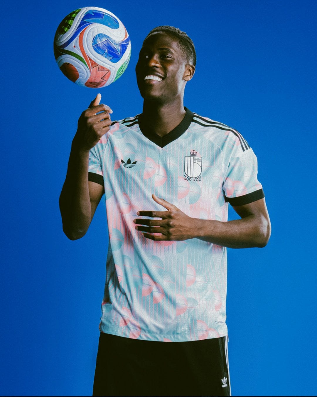 A Belgian soccer player wearing the 2026 Adidas away jersey in pale blue with pink and white accents, smiling while balancing a soccer ball against a bright blue background.