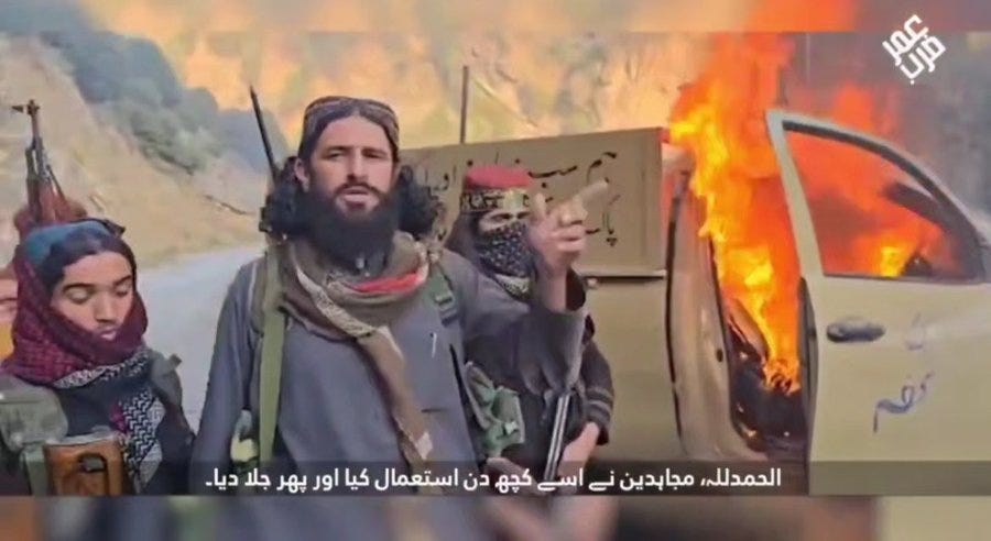 Three armed individuals in traditional attire and headscarves stand near a burning vehicle in a mountainous area; one points forward holding a weapon, another holds a rifle, and a woman stands nearby; flames and smoke rise from the vehicle door; Arabic text overlay appears at the bottom.