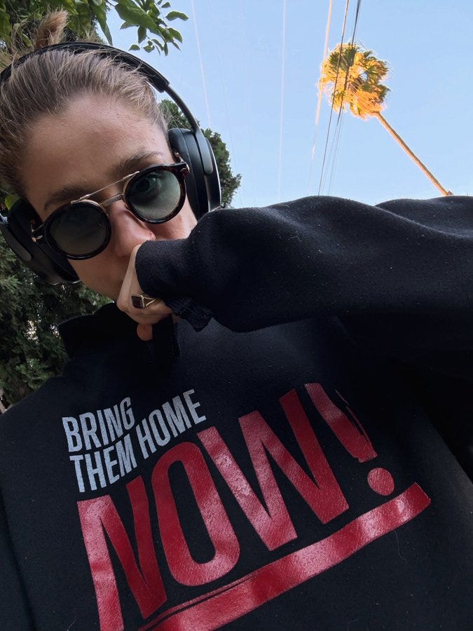 Woman with blonde hair in ponytail wears round black-framed sunglasses and over-ear headphones, posing with hand near mouth in a black hoodie shirt printed with Bring Them Home Now Now Now in bold red and white capital letters, outdoors under blue sky with green palm trees and utility pole in background.