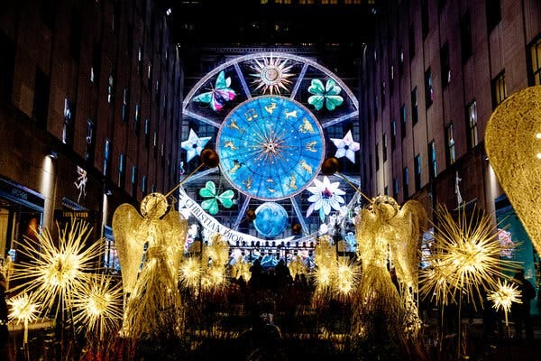 An image of a round blue clock lit up between two buildings in Rockafeller Center. 