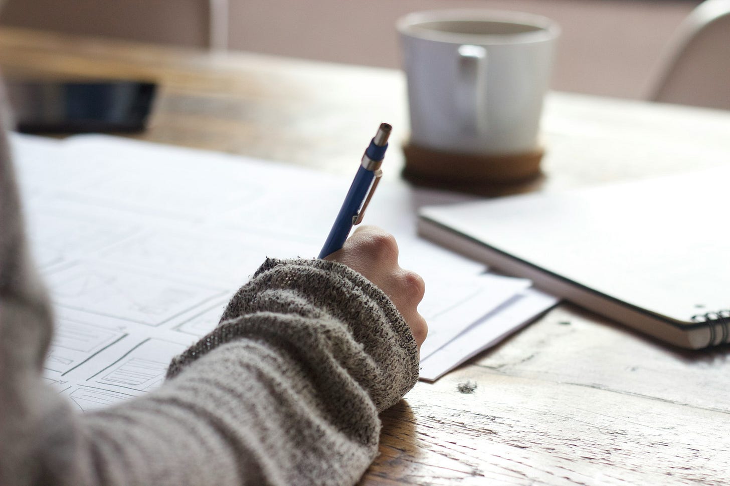 person writing on brown wooden table near white ceramic mug person writing on brown wooden table near white ceramic mug