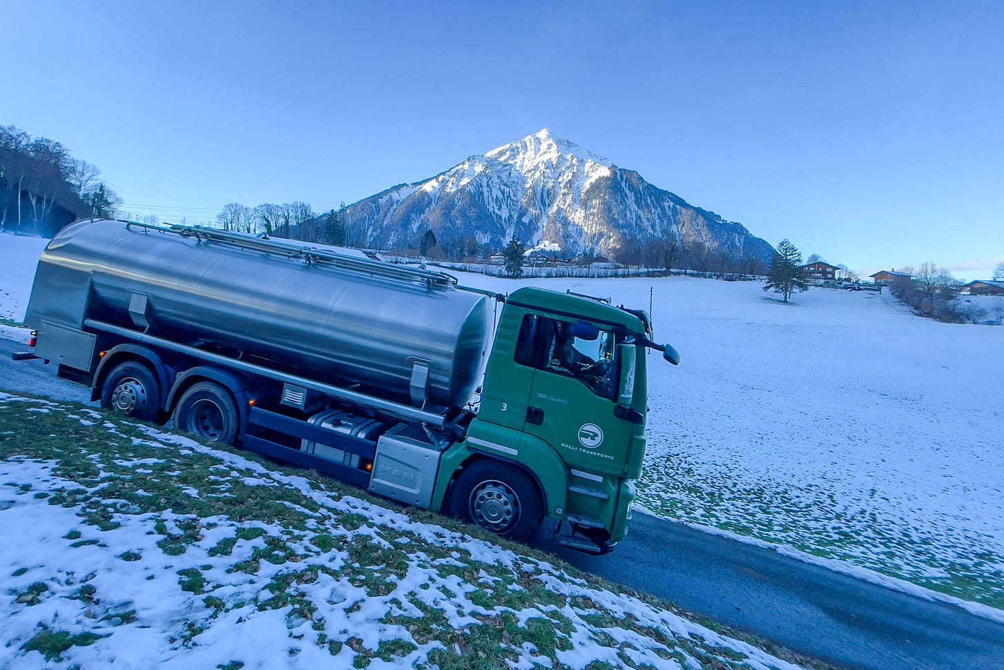 Das Foto zeigt einen grünen Milchsammelwagen der Firma Rolli Transporte, der auf einer vereisten und leicht verschneiten Straße fährt. Der Wagen hat einen silbernen Edelstahltank und ist vor einer malerischen Winterlandschaft positioniert. Im Hintergrund erhebt sich majestätisch der schneebedeckte Gipfel des Niesen, eingerahmt von blauem Himmel. Die umliegenden Wiesen und vereinzelten Bäume sowie ein paar Berghäuser unterstreichen die ländliche Atmosphäre. Das Bild vermittelt einen Eindruck von der täglichen Arbeit in der Milchwirtschaft in den Schweizer Alpen. Das Foto zeigt einen grünen Milchsammelwagen der Firma Rolli Transporte, der auf einer vereisten und leicht verschneiten Straße fährt. Der Wagen hat einen silbernen Edelstahltank und ist vor einer malerischen Winterlandschaft positioniert. Im Hintergrund erhebt sich majestätisch der schneebedeckte Gipfel des Niesen, eingerahmt von blauem Himmel. Die umliegenden Wiesen und vereinzelten Bäume sowie ein paar Berghäuser unterstreichen die ländliche Atmosphäre. Das Bild vermittelt einen Eindruck von der täglichen Arbeit in der Milchwirtschaft in den Schweizer Alpen.