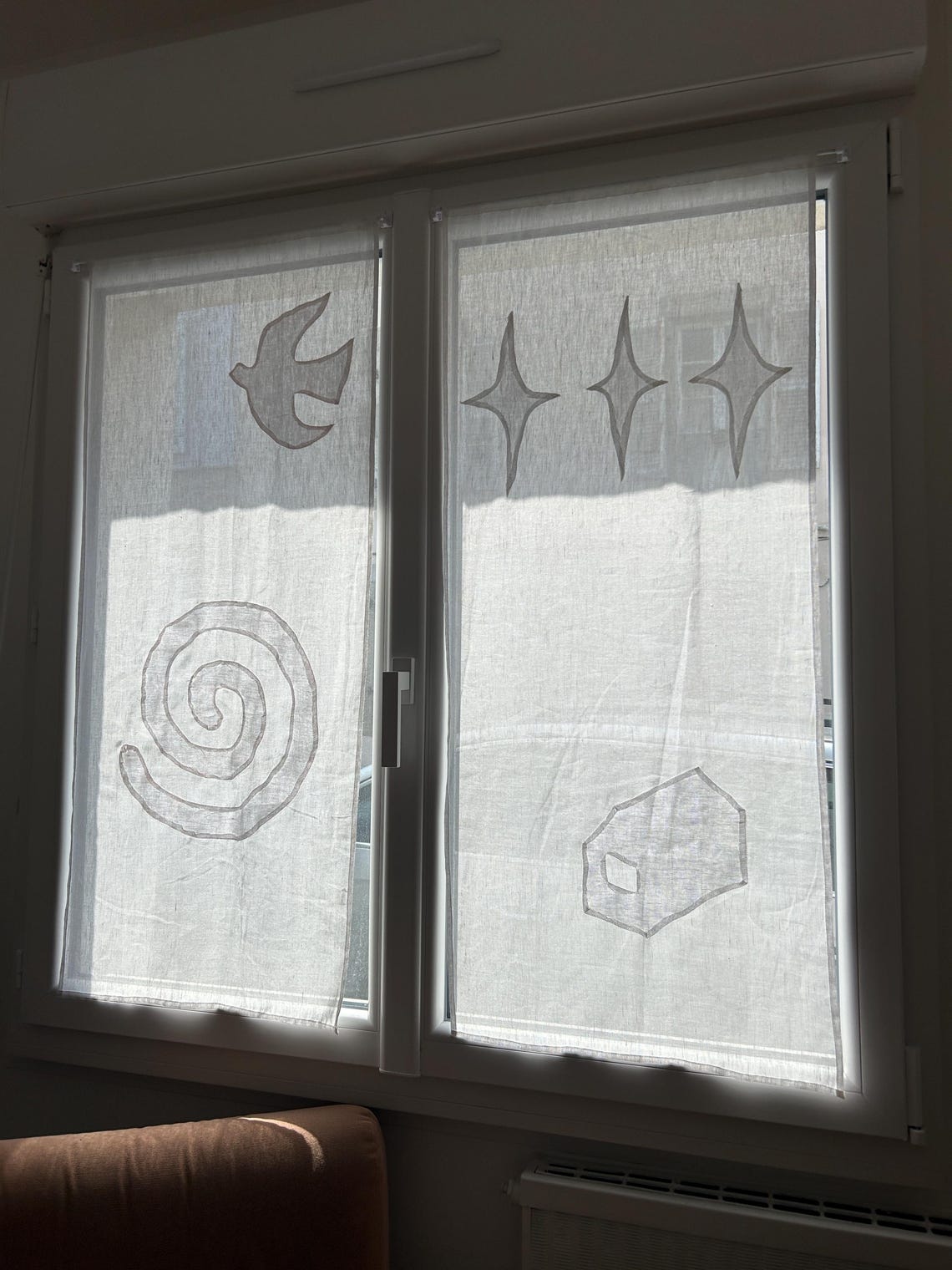 May include: Sheer white window curtains with hand-drawn gray designs. The designs include a bird, a spiral, three starbursts, and a house. The curtains are hanging in a white window frame, allowing sunlight to filter through.