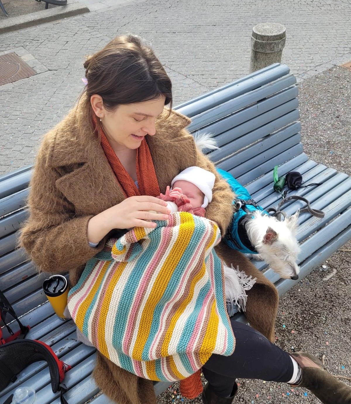 A new mother bottle-feeding her child on a bench, looking tired, with a Jack Russell mix next to her