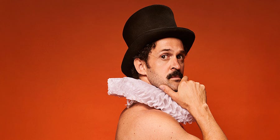 Performer Damien Warren-Smith as Garry Starr, posing shirtless in a dramatic side profile against a red backdrop, wearing a black top hat and a white Elizabethan ruff collar.