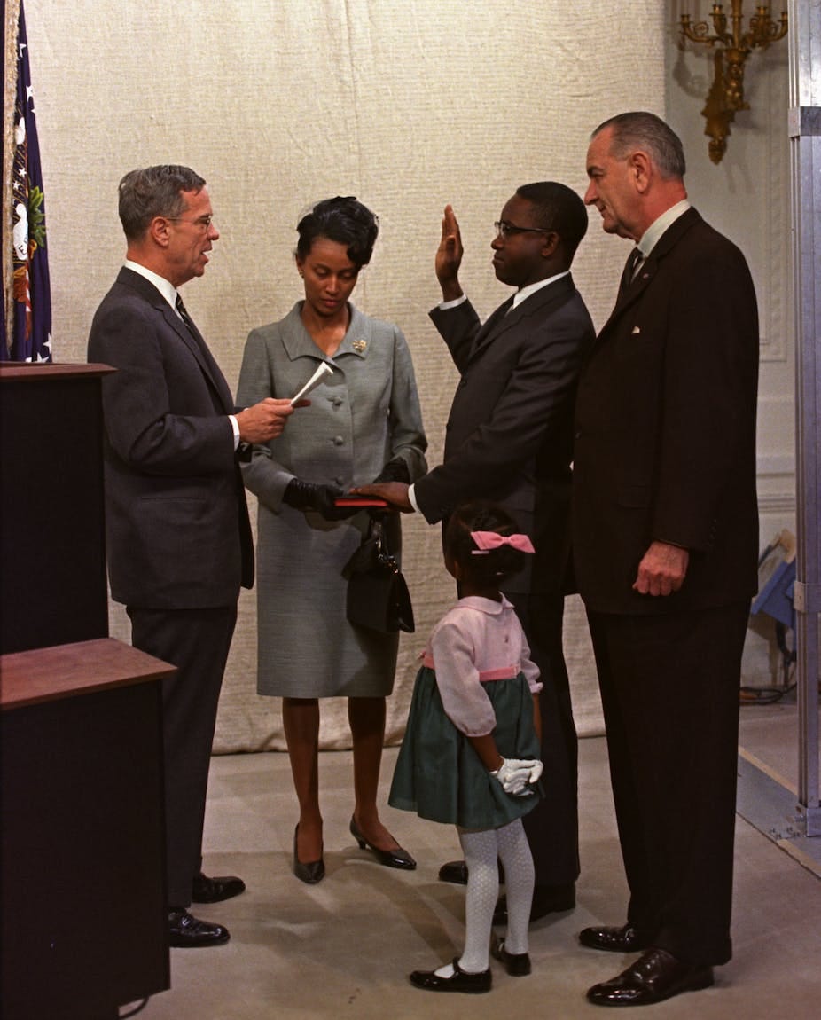 A man with his left hand on a bible held by a woman on his right, holds his right hand face-high, palm side out, as he speaks to a man facing him. Another man and little little girl stand close by on his left. A man with his left hand on a bible held by a woman on his right, holds his right hand face-high, palm side out, as he speaks to a man facing him. Another man and little little girl stand close by on his left.