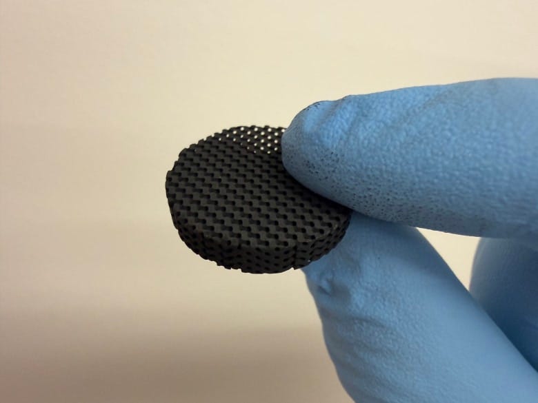Hand in blue glove holding a circular, 3D-printed battery component with a grid-like surface. Hand in blue glove holding a circular, 3D-printed battery component with a grid-like surface.