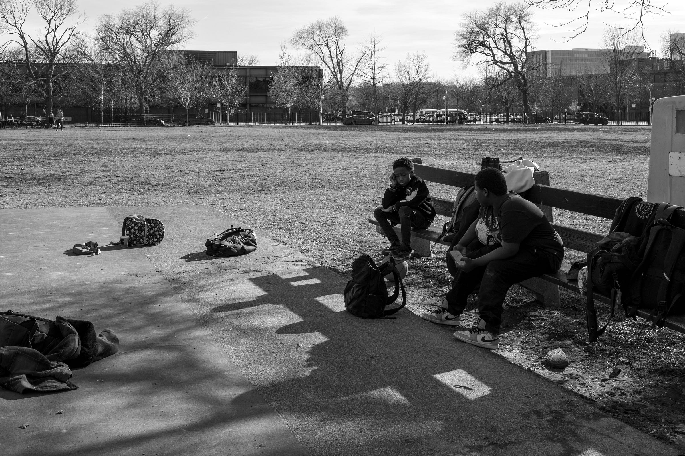 A black-and-white photograph of two young boys sitting on a park bench, surrounded by scattered backpacks and personal belongings. One boy rests his head on his hand, gazing into the distance, while the other eats a snack, engaged in conversation. Shadows from the bench and surrounding objects stretch across the pavement. In the background, an open field, trees, and a row of parked cars set the urban scene, creating a quiet moment of reflection and companionship.