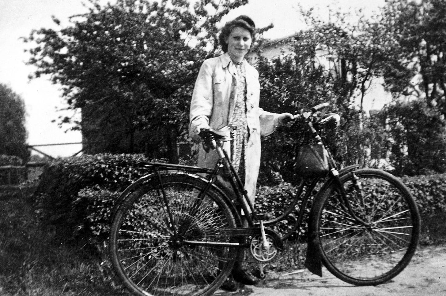 Young woman in a coat and a flower dress with an old bike in front of a hedge, black and white photo 