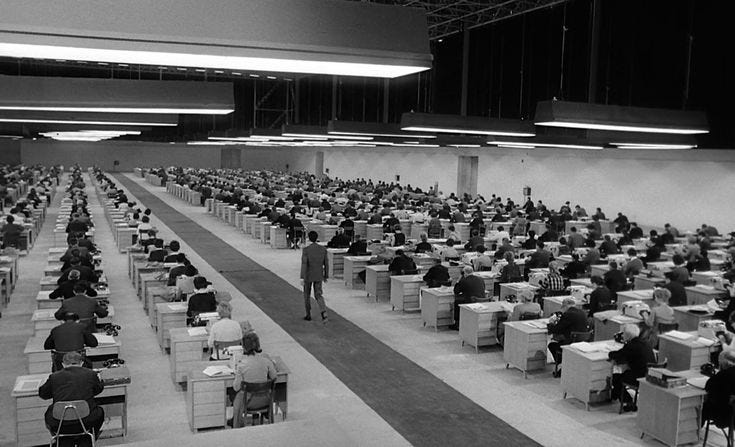 This may contain: many people are sitting at desks in an empty room with lots of computers on them