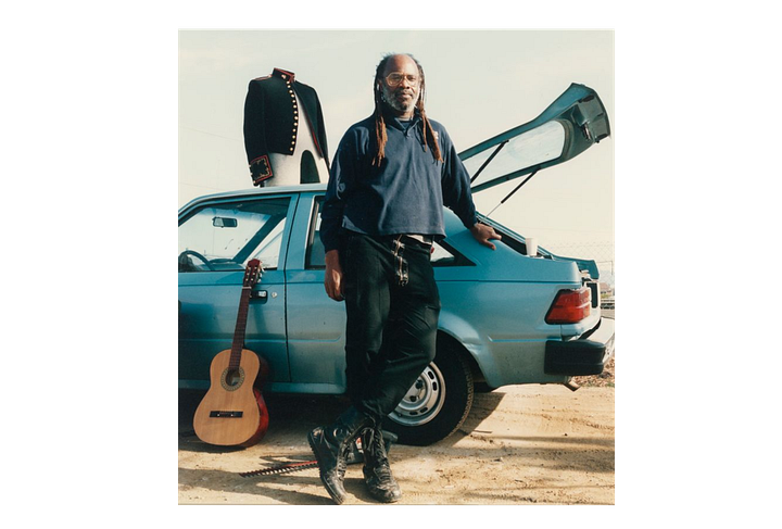 A girl in a vintage truck with vintage dolls, two men sitting on a car, one man with dreadlocks in front of a hatchback and a guitar