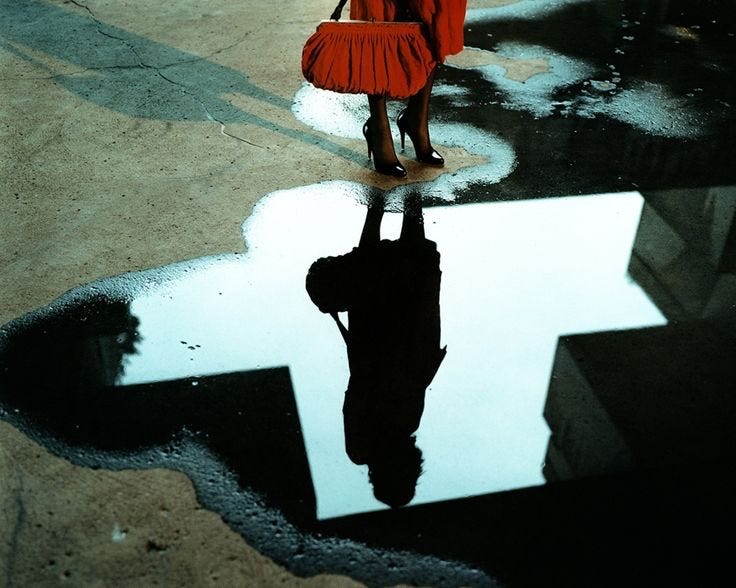 This may contain: a woman in a red dress is holding an umbrella and standing on the street with her reflection