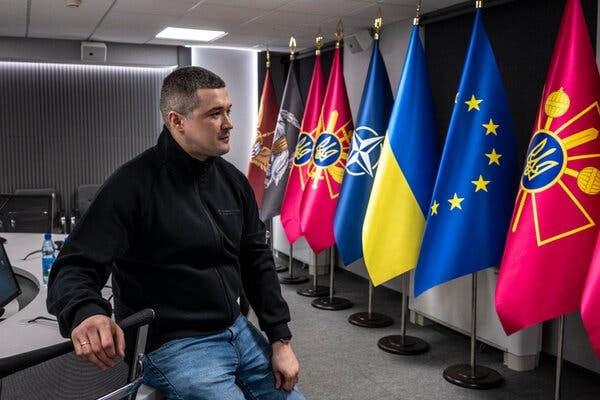 A person wearing a black jacket and bluejeans sits on a conference room table. In the background, a line of flags on poles displays blue and yellow, blue with yellow stars, and various red, blue, and dark gray designs.