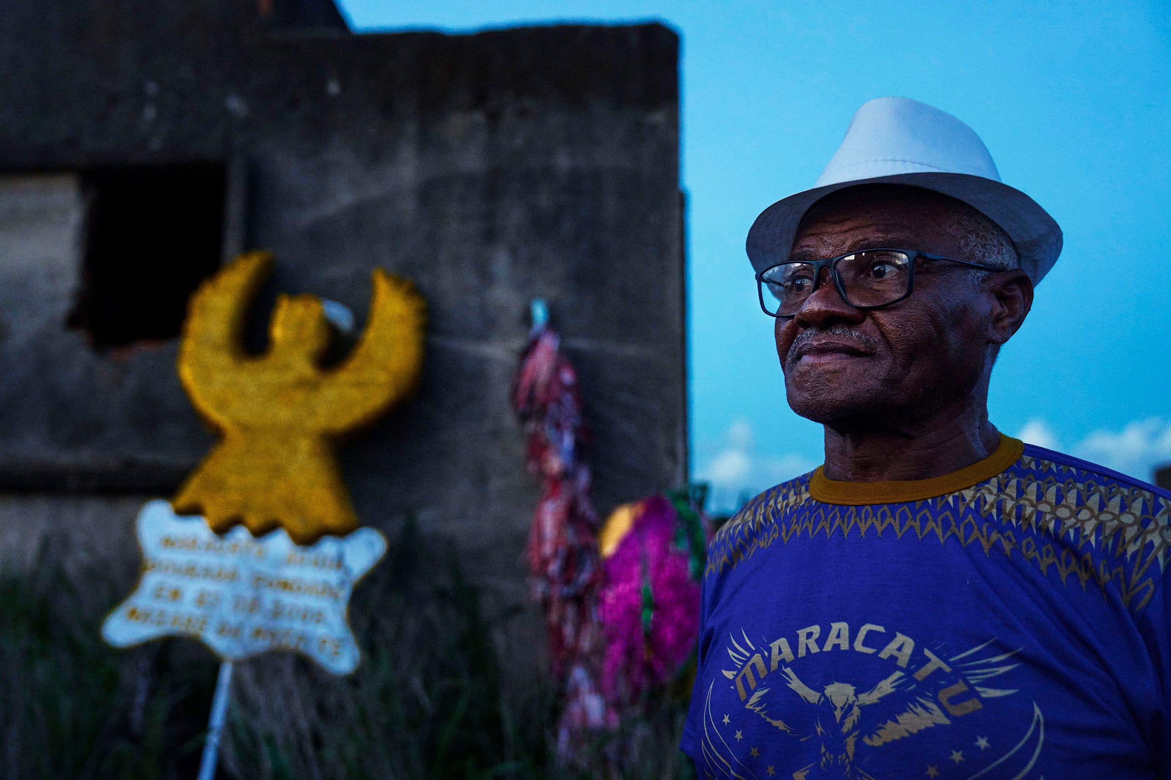 An older man in glasses and a white hat stands outside near a concrete structure. Behind him are colorful wreaths, including a yellow angel, and a sign in Portuguese. The sky is blue and it appears to be early evening.