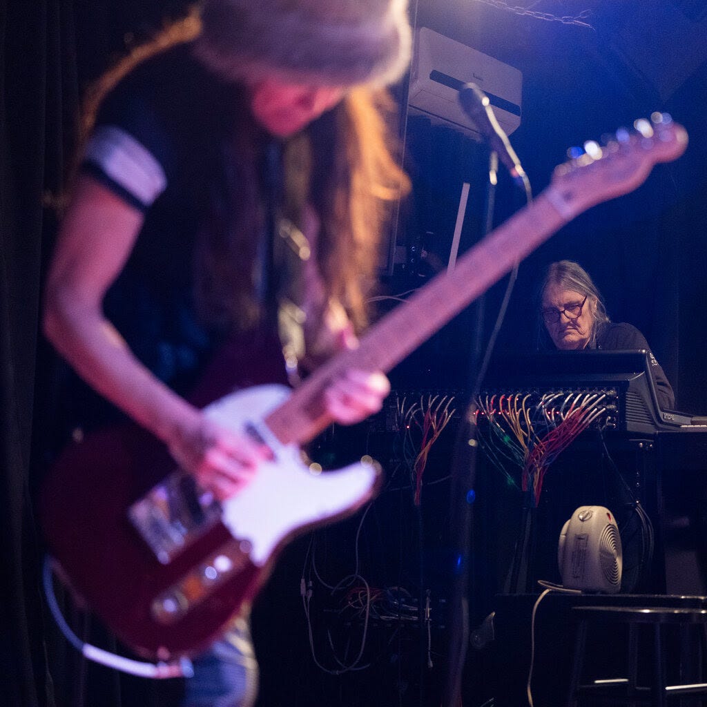 A guitarist in the foreground plays an electric guitar while in the back, a man with gray hair looks down studiously at a sound board.
