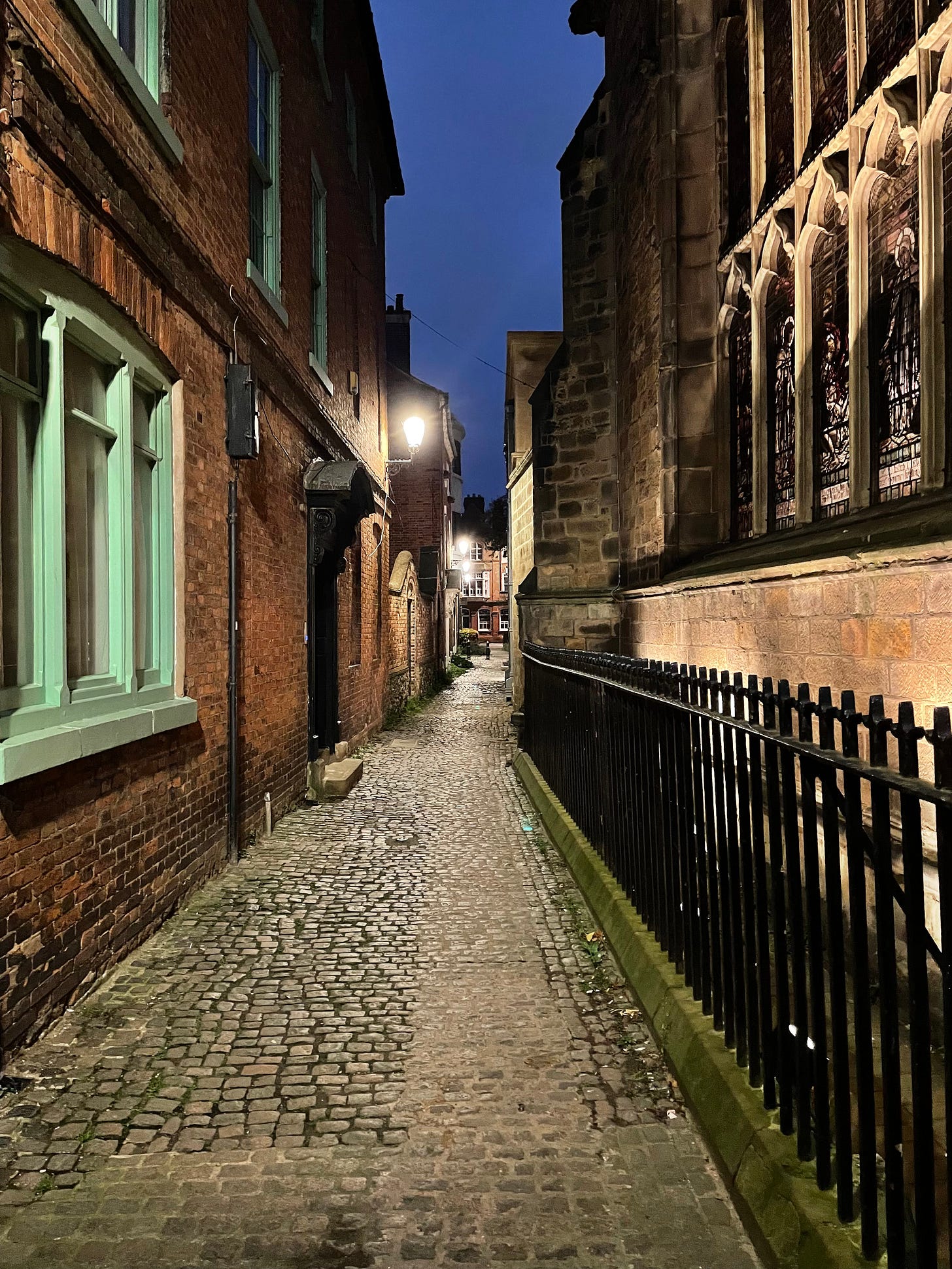A narrow old-fashioned lane, with the elaborate stained glass window of the cathedral glowing in the dark on the right hand side, and an old brick building lit by a Narnia-esque street lamp on the left.