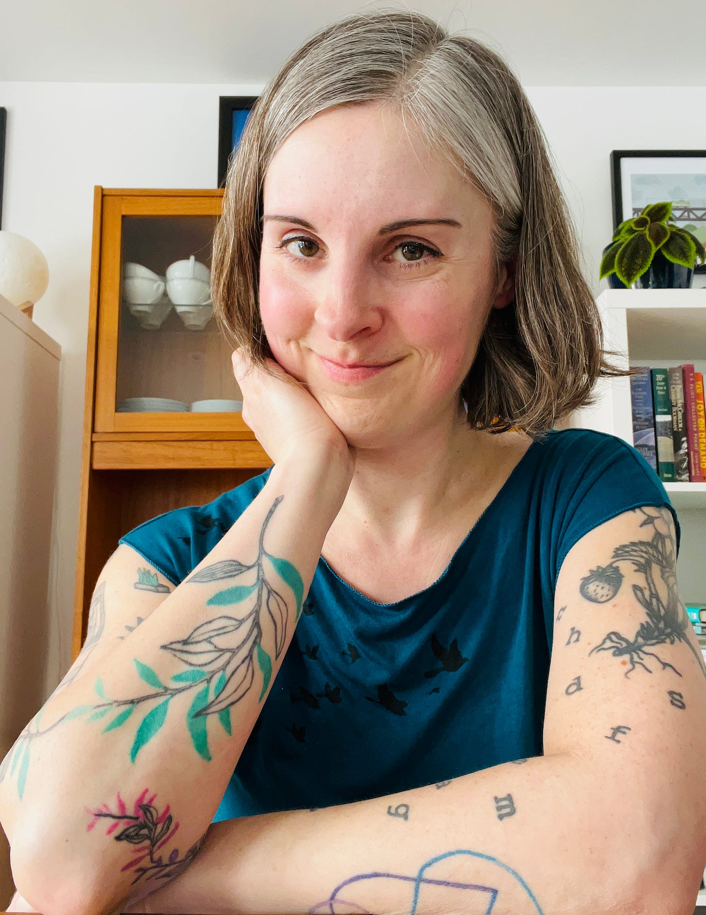 Selfie of Jen in her dining room, sitting in front of a dish cabinet and bookshelves, wearing a blue shirt and looking somewhat authorly.