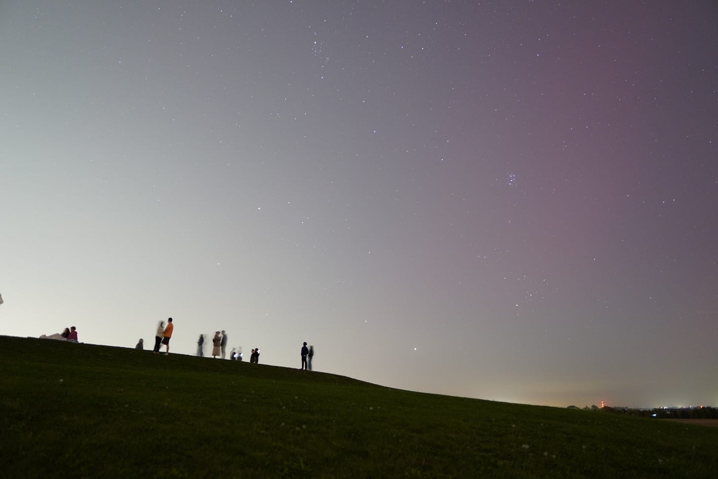 A silhouette of a hill with about a dozen people on it on the left and city lights on the right. In the top right is a patch of red-tinged sky as opposed to the rest of the gray sky.