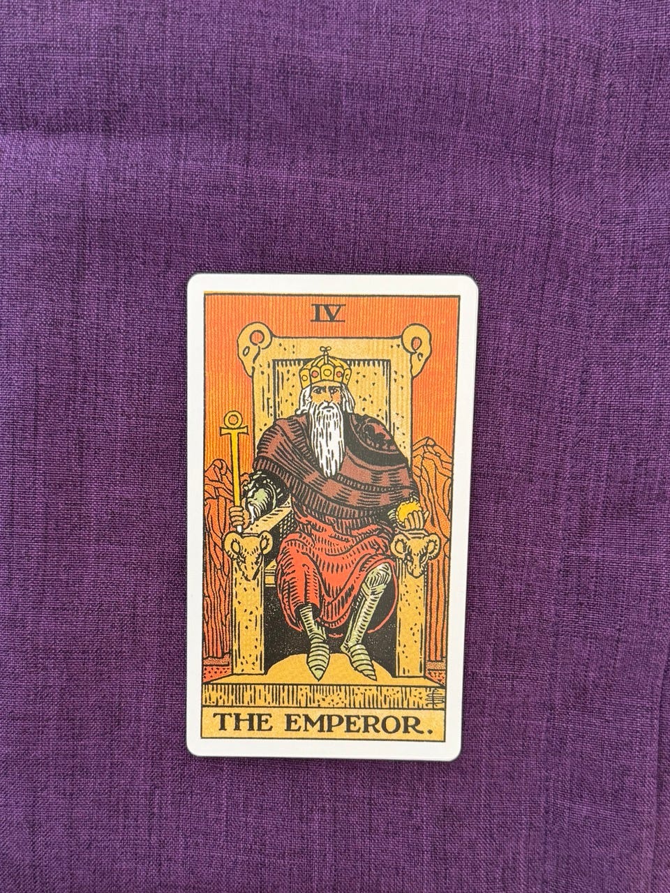 An old man, with long white beard sits upon a large stone throne, adorned with four rams’ heads. In his right hand, The Emperor holds an ankh (handle shaped cross) and in his left is an orb. He wears a red robe, underneath it, he wears a suit of armor and a golden crown. Behind his throne looms a tall, impenetrable mountain range, beneath the peaks flows a small river.