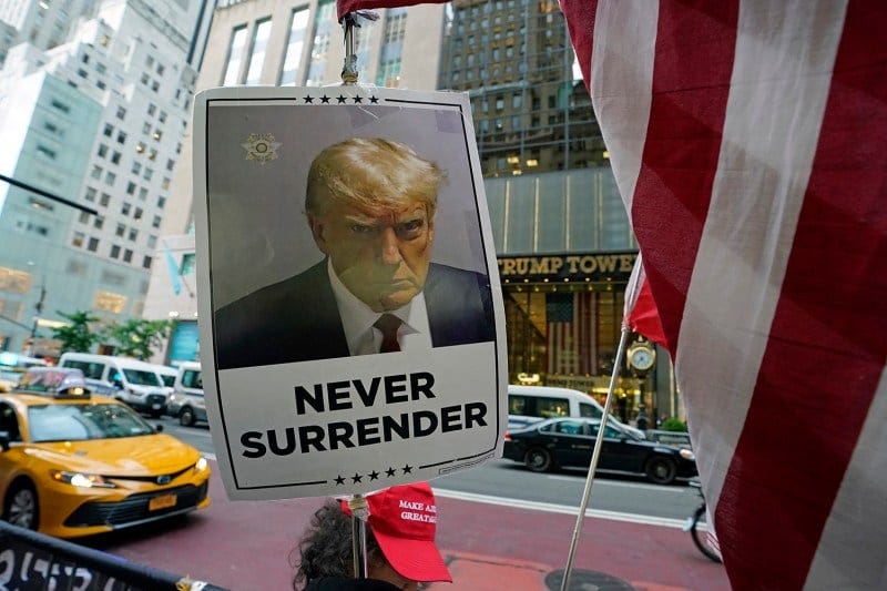 A protester wearing a Make America Great hat holds a sign with a mugshot of Donald Trump on it and the words "Never Surrender." A taxi, other cars, Trump Tower, and a U.S. flag are seen behind the sign. A protester wearing a Make America Great hat holds a sign with a mugshot of Donald Trump on it and the words "Never Surrender." A taxi, other cars, Trump Tower, and a U.S. flag are seen behind the sign.