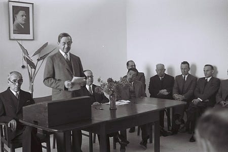 Black and white photograph of a suited man reading a speech. The man stands in front of a table. Seven men in suits sit behind him.