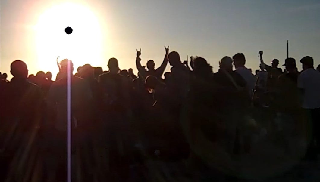 the black sun beaming across a group of people watching a drum circle perform on a beach