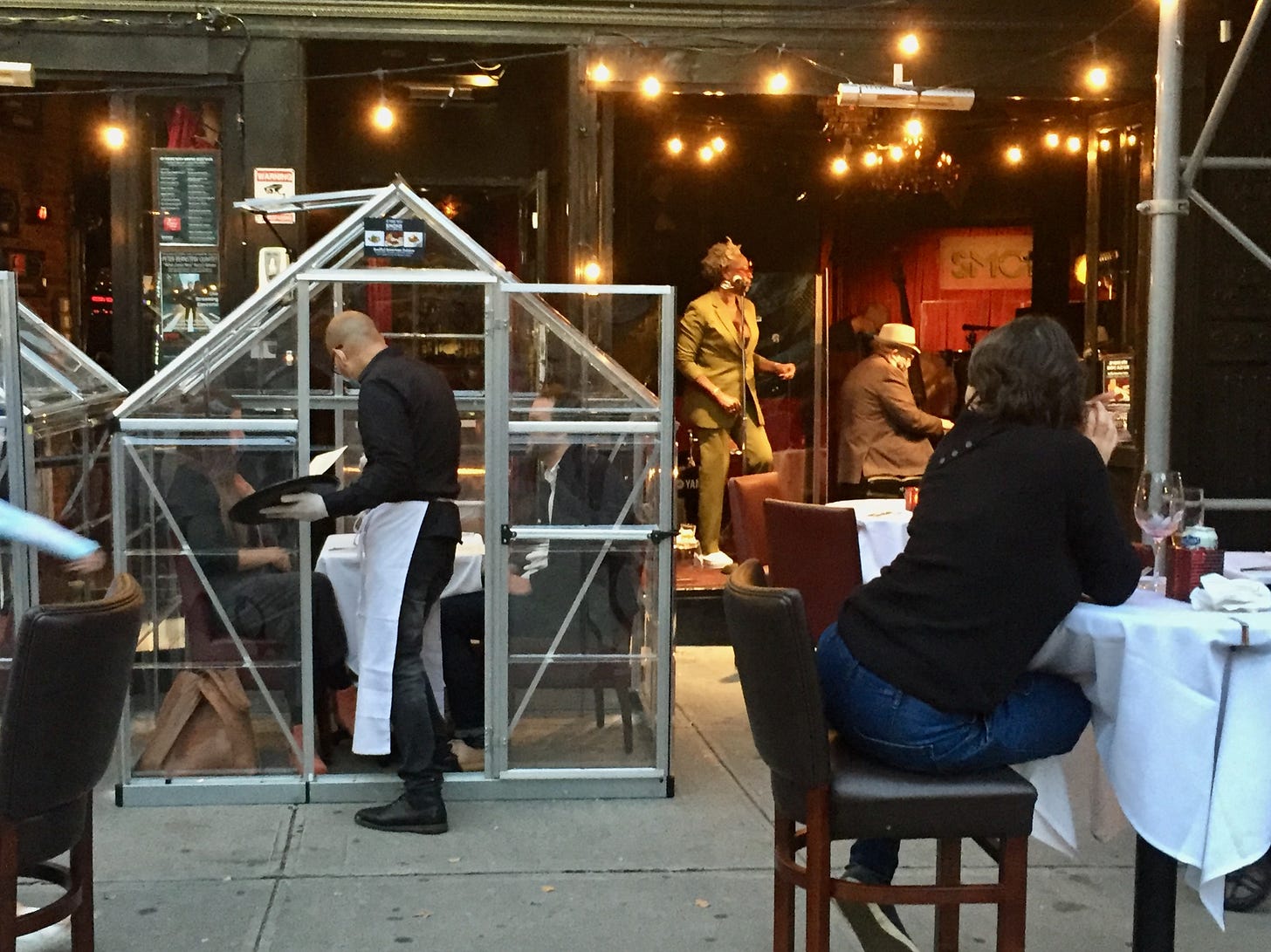 A waiter serves a customer in a Covid-era enclosure on a sidewalk while a jazz singer croons in the background