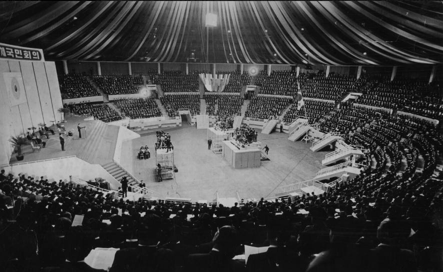 Wide interior view of Jangchung Gymnasium filled with thousands of delegates in uniform rows. A central stage and ballot box are visible under a canopy of overhead drapery, with media crews documenting the event.