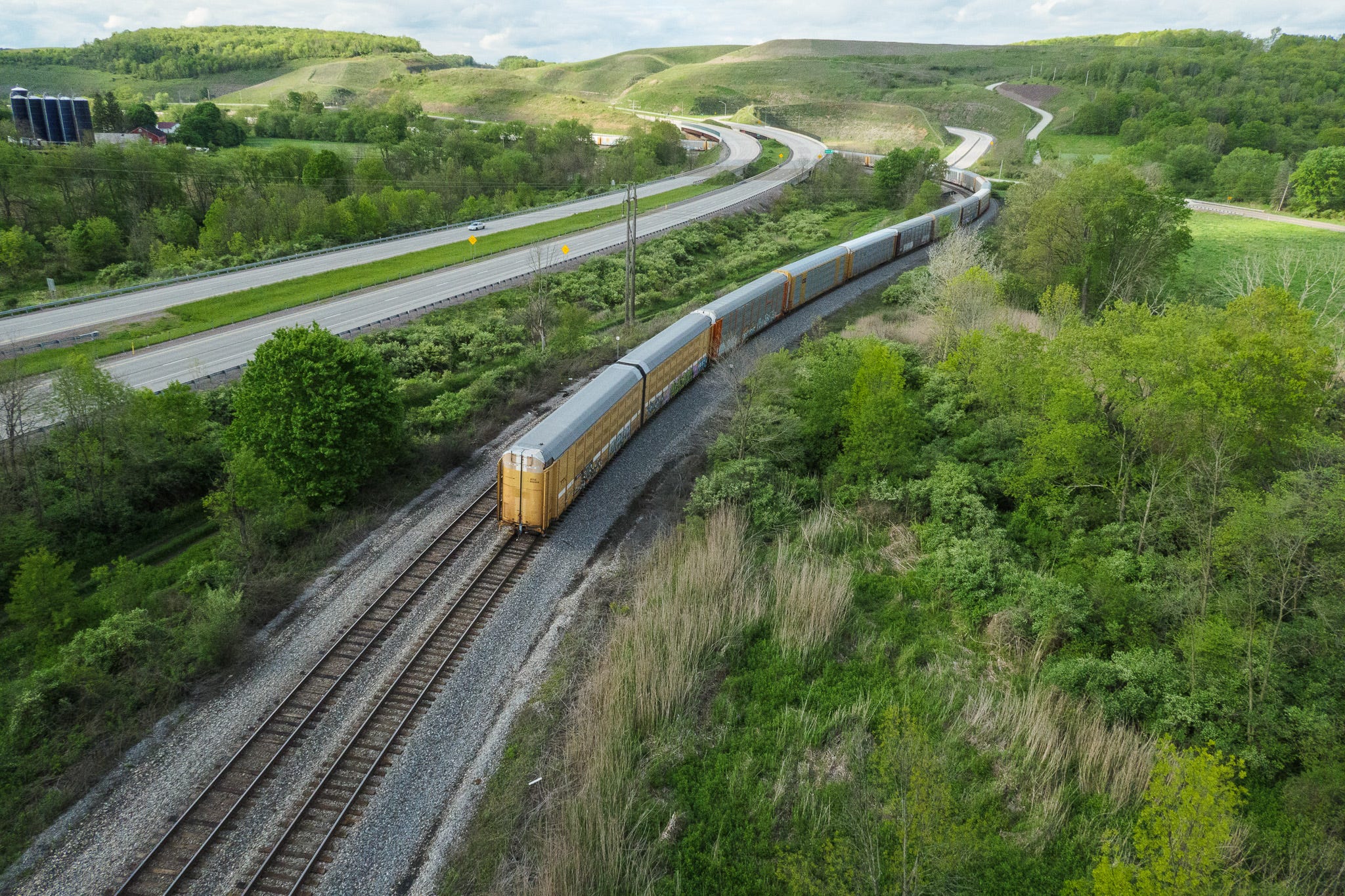 Photo looking down upon a train as it runs along a curving train track. The front of the train cannot be seen in the distance beyond the curve. The rear of the train features two cars painted yellow. I modern four-lane highway runs alongside the railroad track and bends to the left also. In the distance, rolling hills.